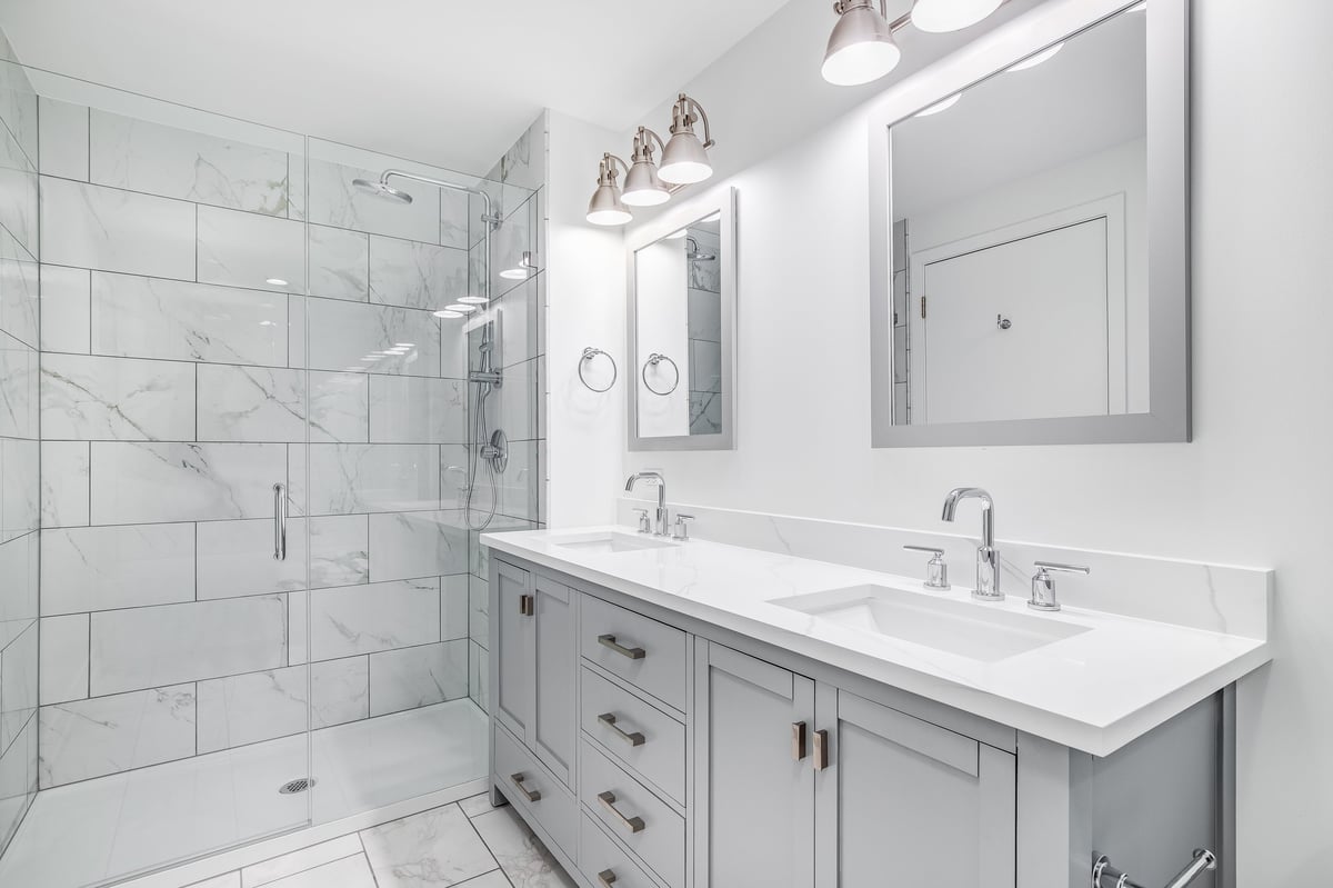 Remodeled bathroom with a grey vanity, bronze hardware, marble tile, and a glass shower enclosure