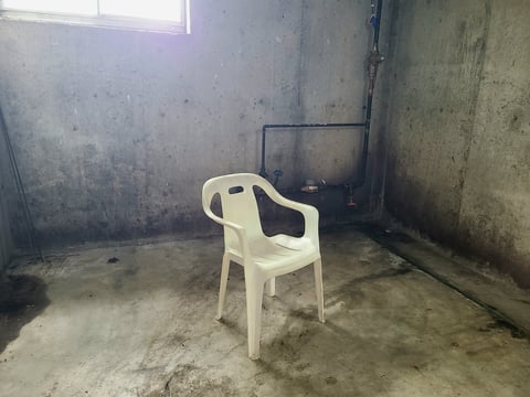 A dirty, dingy basement with a window as the only lighting. Residential pipes can be seen in the background. A white plastic chair sits in the center of the room, creating an unsettling atmosphere.