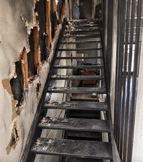 Narrow staircase with deteriorating walls, showing peeling paint and damage on the left side, leading downward between metal railings