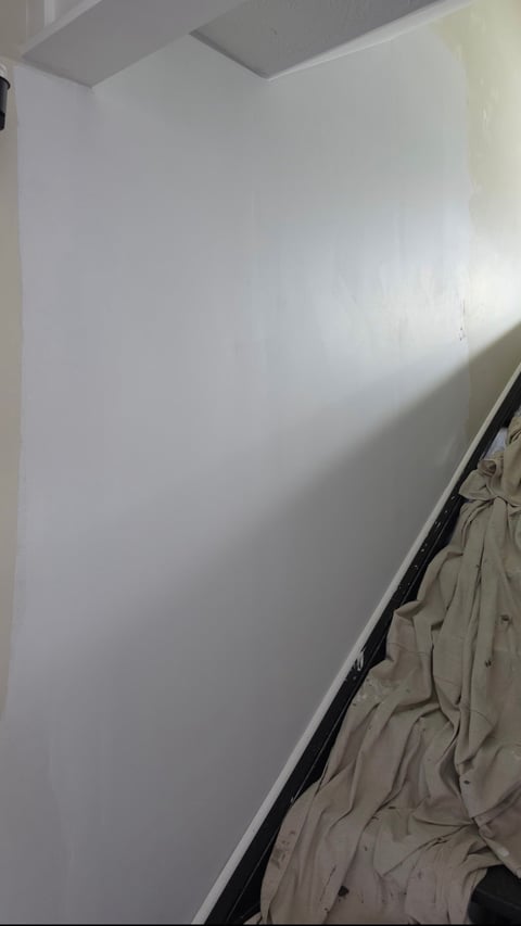 Empty white bedroom wall with sunlight streaming across it and beige bedding visible at the bottom right corner