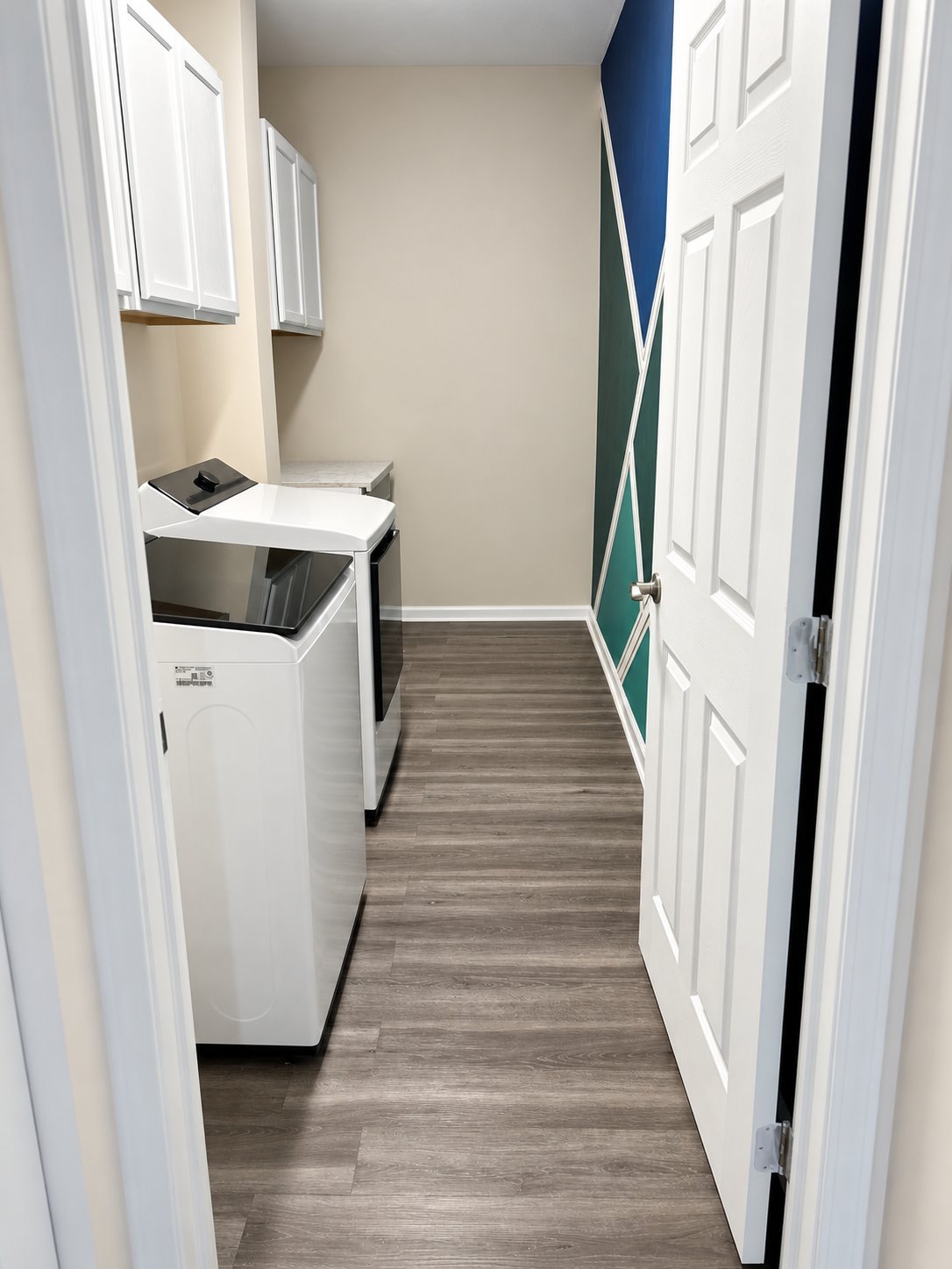 Doorway view of Arlene H.’s new laundry room, showing white appliances and cabinetry, gray wood-look flooring, and a blue-and-green geometric accent wall.