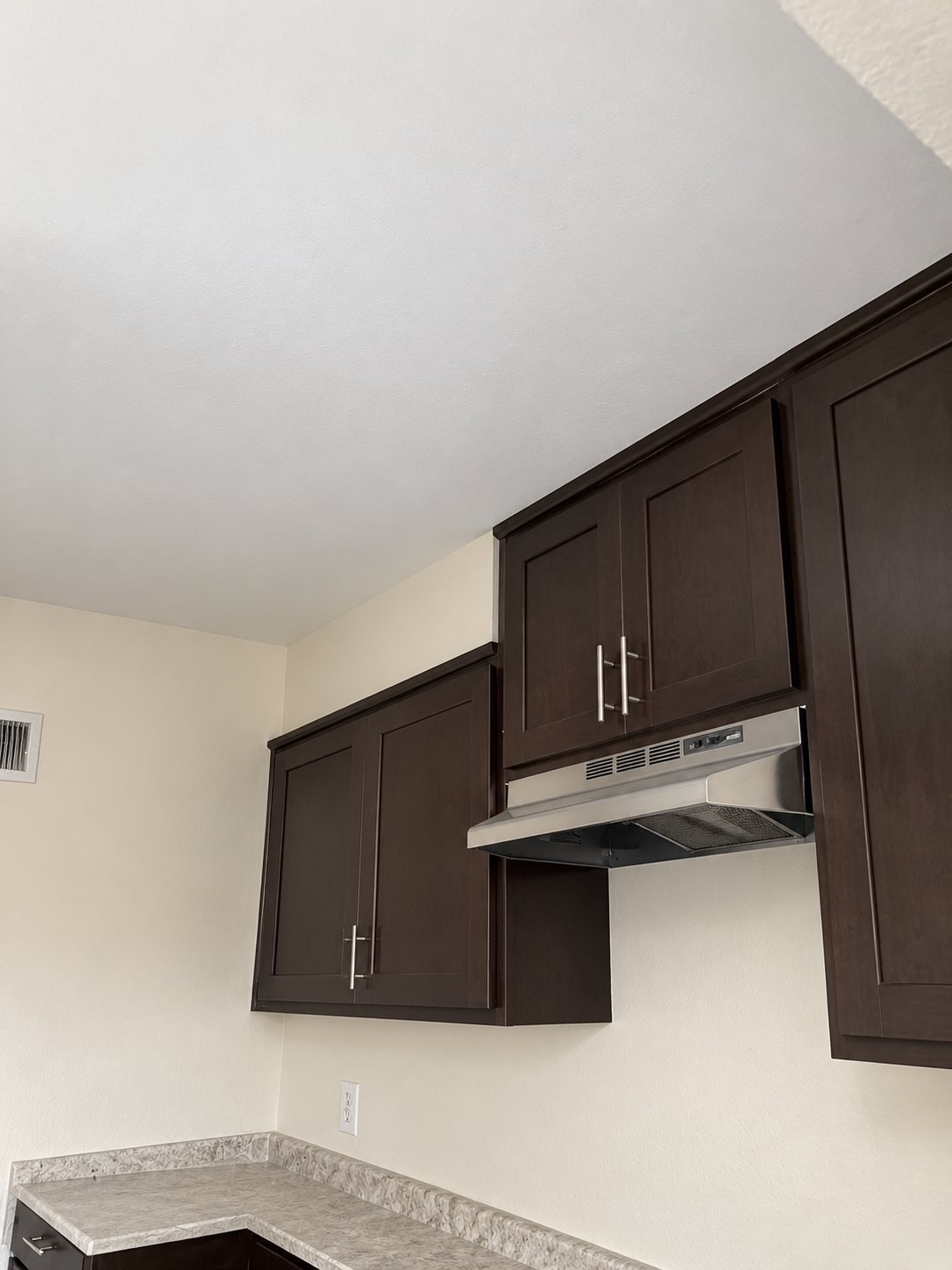 Dark brown kitchen cabinets with stainless steel range hood mounted on cream-colored wall above granite countertop