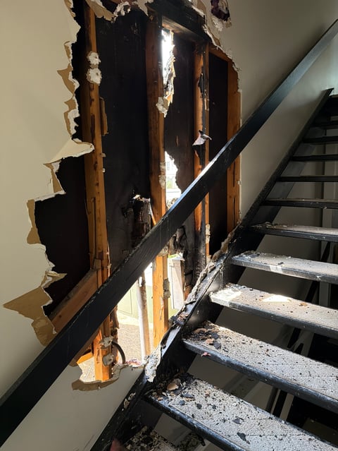 Damaged interior wall showing exposed wooden studs, drywall, and black metal staircase railing with visible deterioration and debris