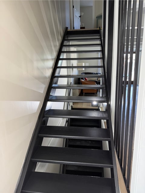 Black metal staircase with glass panels ascending between white walls and vertical blinds in modern interior